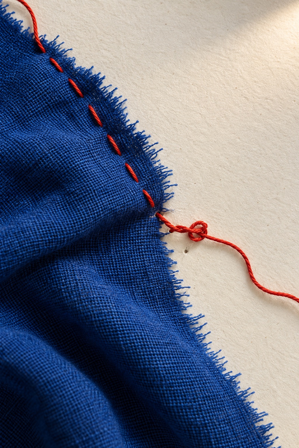A red thread stitches blue fabric to ivory paper, connecting the two with a loose knot.
